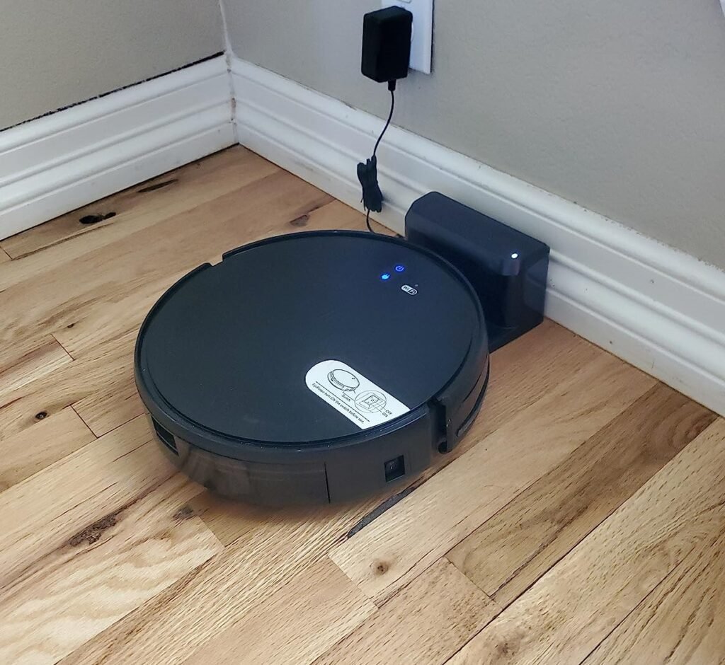 A black robot vacuum with mop function docking and charging at a charging station in a corner of a hardwood floor room.