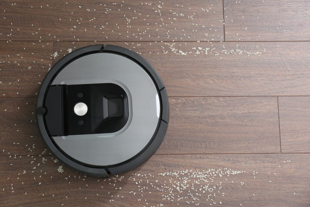 A robot vacuum cleaner navigating across a hardwood floor, picking up scattered rice grains.