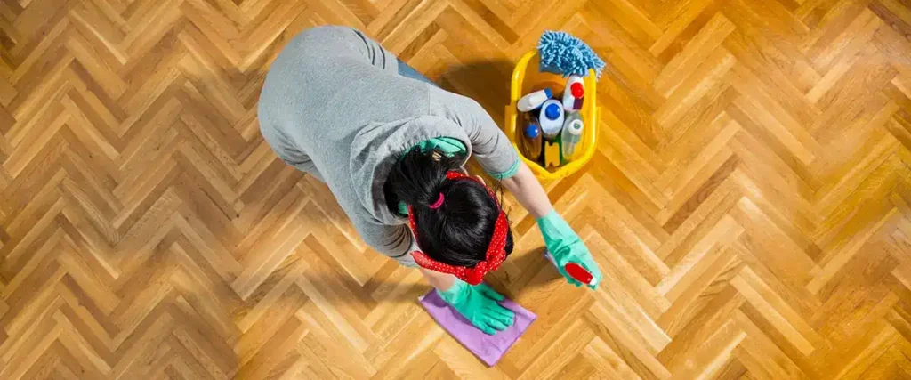 A woman wearing gloves and a headband maintains a beautifully restored parquet floor by cleaning it with a cloth. A bucket filled with cleaning supplies is placed nearby, showcasing the regular maintenance required to keep parquet flooring in pristine condition.