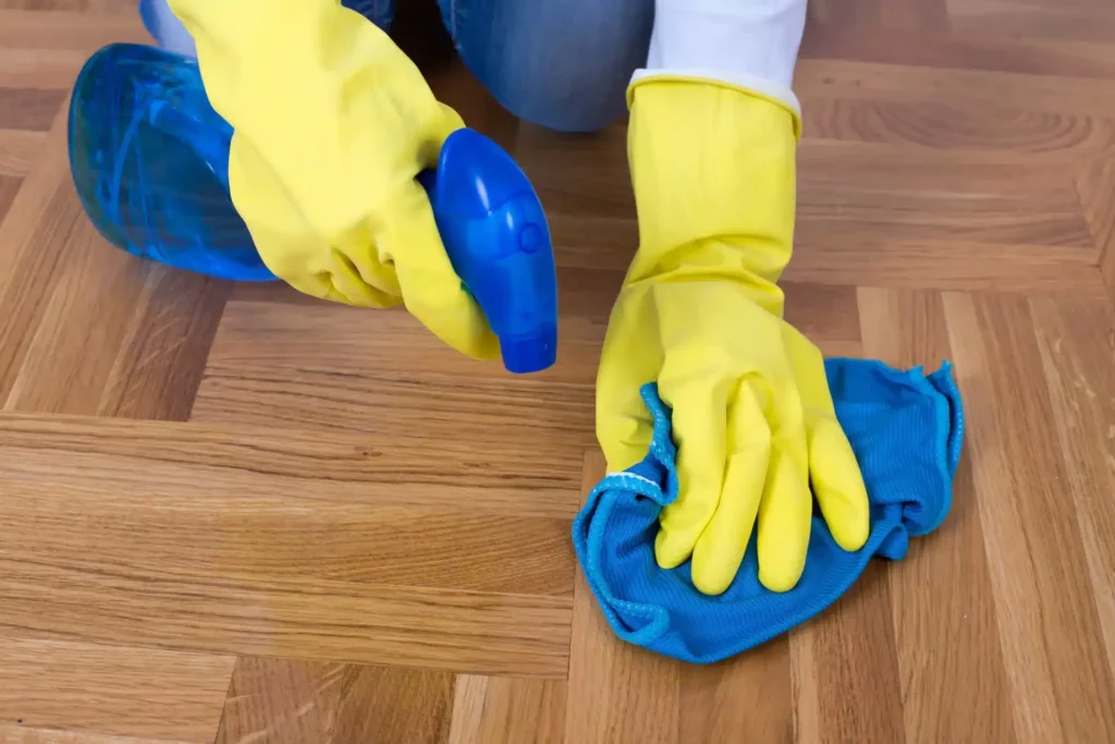 A person wearing yellow gloves is cleaning a newly refinished wooden floor with a blue cloth and a spray bottle. The herringbone parquet floor has a smooth, glossy finish, indicating a high-quality sanding and refinishing job by Mr Sander®.